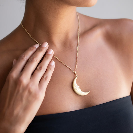 A close-up of a model wearing a delicate gold chain necklace with a crescent moon pendant. The pendant features a small carved face on the side.