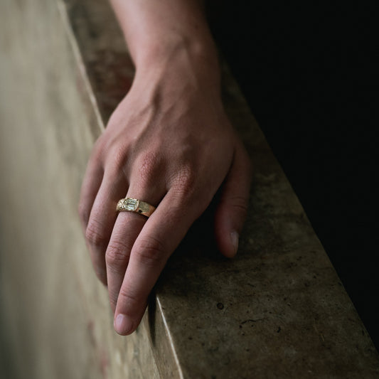A close-up of a person's hand resting on a stone ledge, wearing a gold ring with a rectangular clear gemstone.