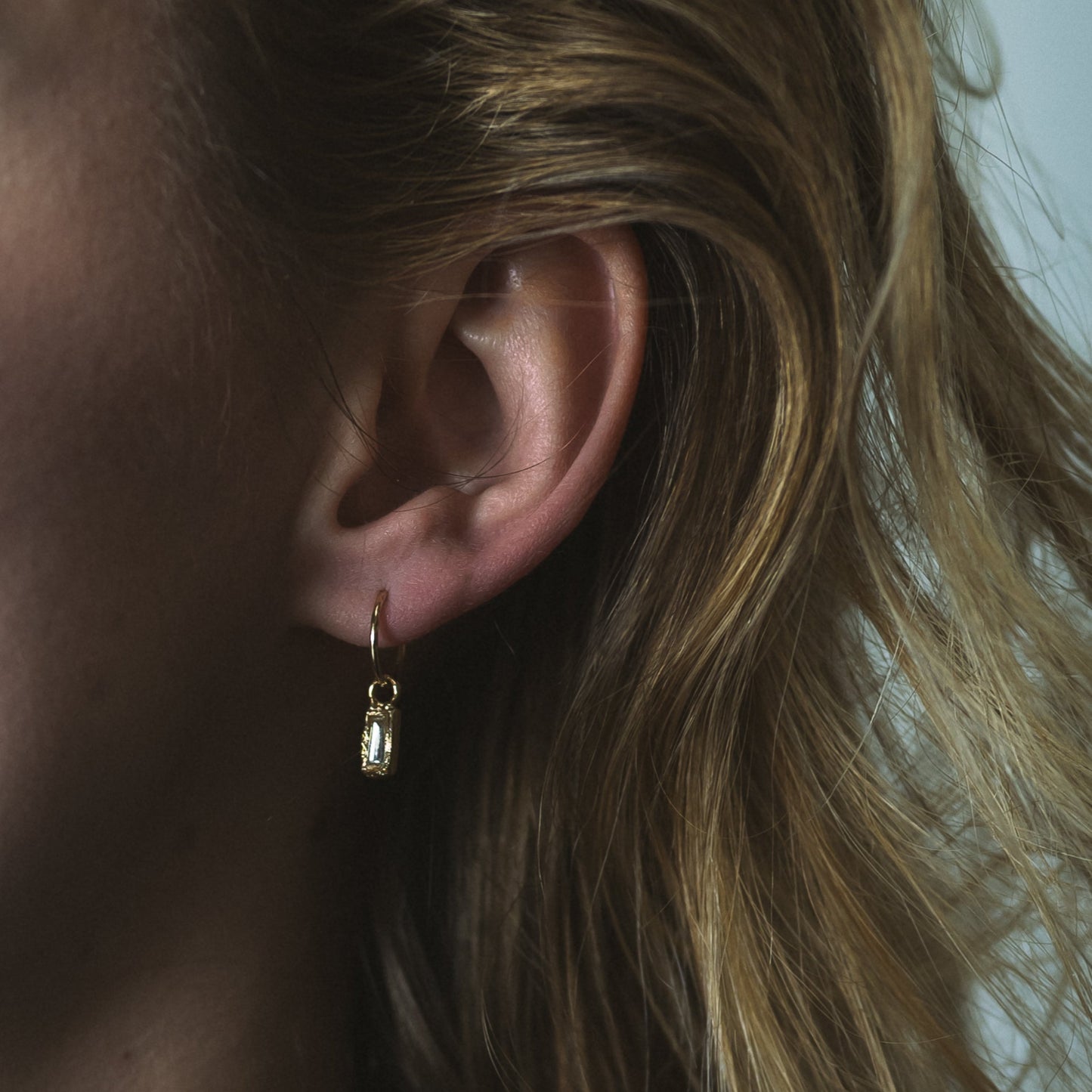 Ellis Mhairi Cameron - Close-up of an ear wearing a gold earring with a small charm, hair partially covering the face.