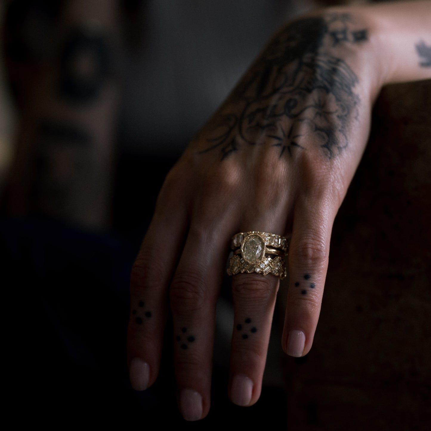 Ellis Mhairi Cameron - Hand with tattoos wearing a gold ring against a dark background