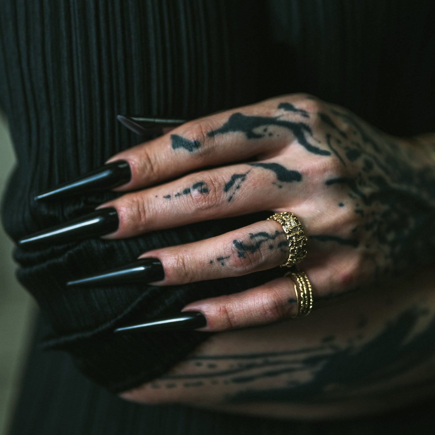 Ellis Mhairi Cameron - Hand with tattoos wearing black nail polish and gold rings on a dark background