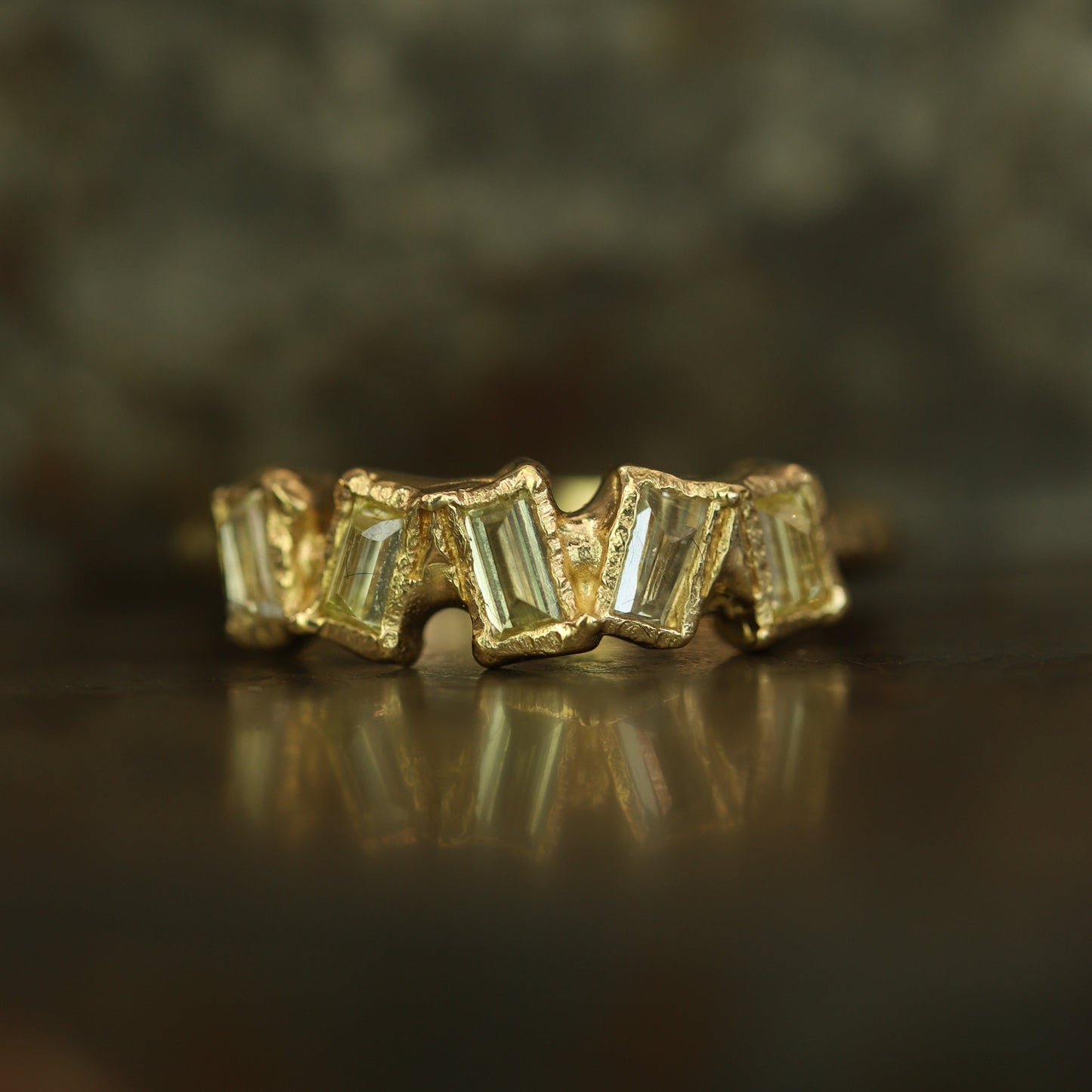 Ellis Mhairi Cameron - Gold ring with baguette-cut gemstones on a reflective surface with a blurred background