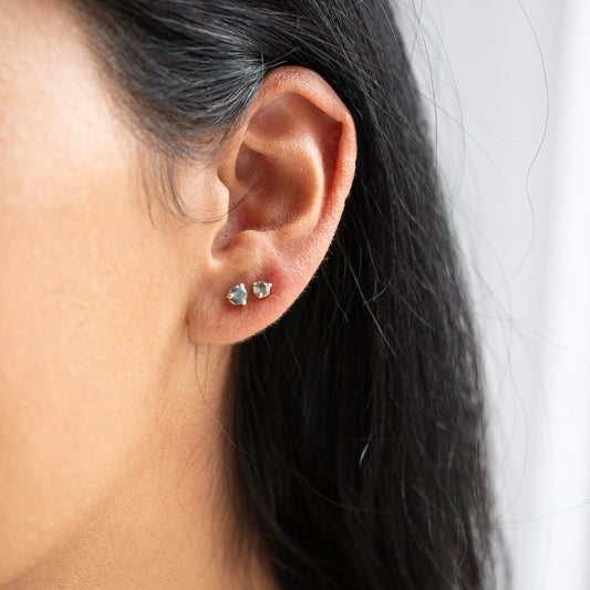 A close-up of a person's ear wearing two small silver stud earrings with blue stones, against a light neutral background.