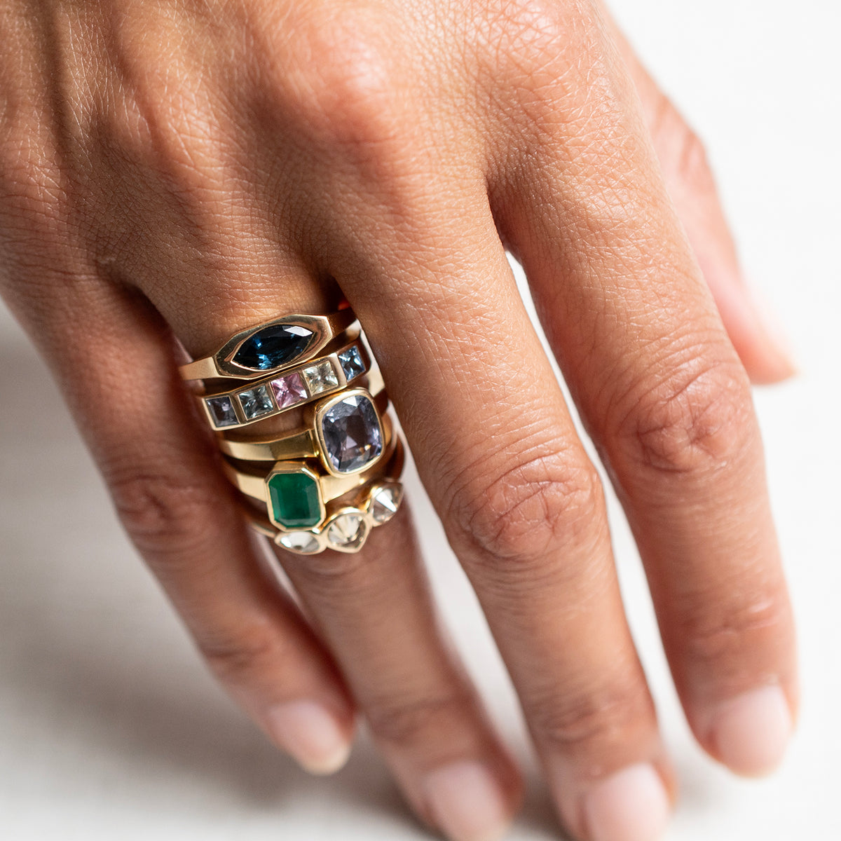 A close-up of a hand wearing a stack of gold rings with various colored gemstones, including blue, green, and clear stones, against a bright white background.