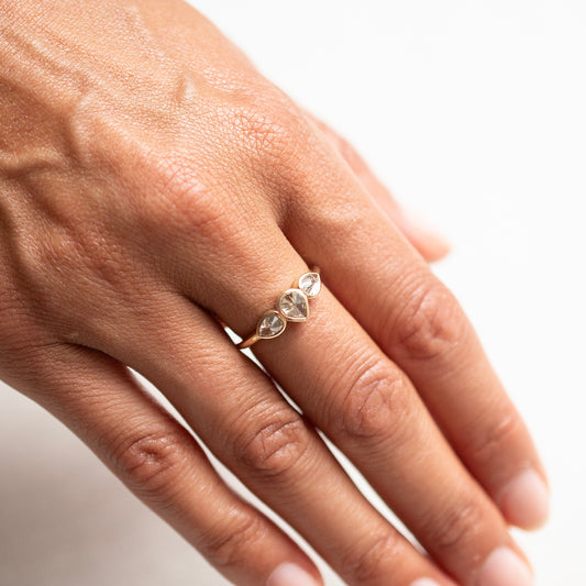 A close-up of a hand wearing a delicate gold ring with three small clear teardrop-cut gemstones, set against a bright white background.