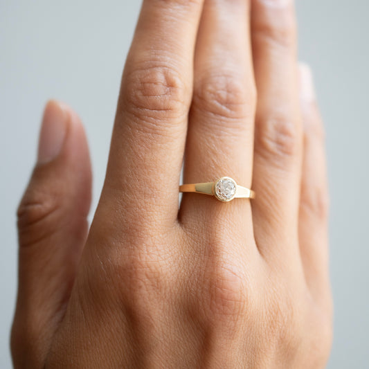 A close-up of a hand wearing a delicate gold ring with a round clear diamond, set against a soft light gray background.