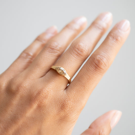 A close-up of a hand wearing a delicate gold ring with a marquise-cut clear gemstone in a bezel setting, photographed against a soft light gray background.