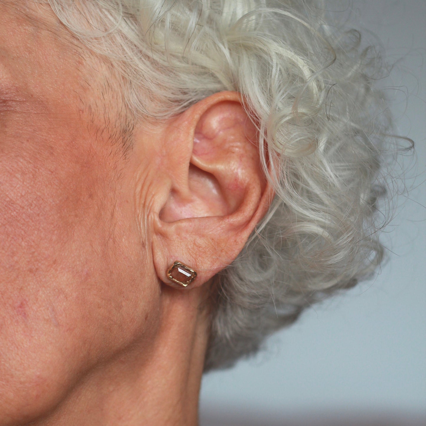 Ellis Mhairi Cameron - Close-up of an older woman's ear with a gold earring against a neutral background