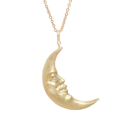 A gold crescent moon pendant necklace with a sculpted face and a small clear diamond in one eye, suspended from a delicate gold chain against a plain white background.
