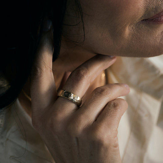 A close-up lifestyle shot of a person wearing a textured gold ring with a dark oval stone on their ring finger, set against a soft cream-colored shirt and warm, dim lighting.