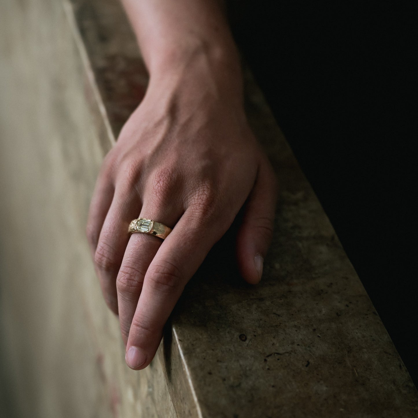 A close-up of a person's hand resting on a stone ledge, wearing a gold ring with a rectangular clear gemstone.
