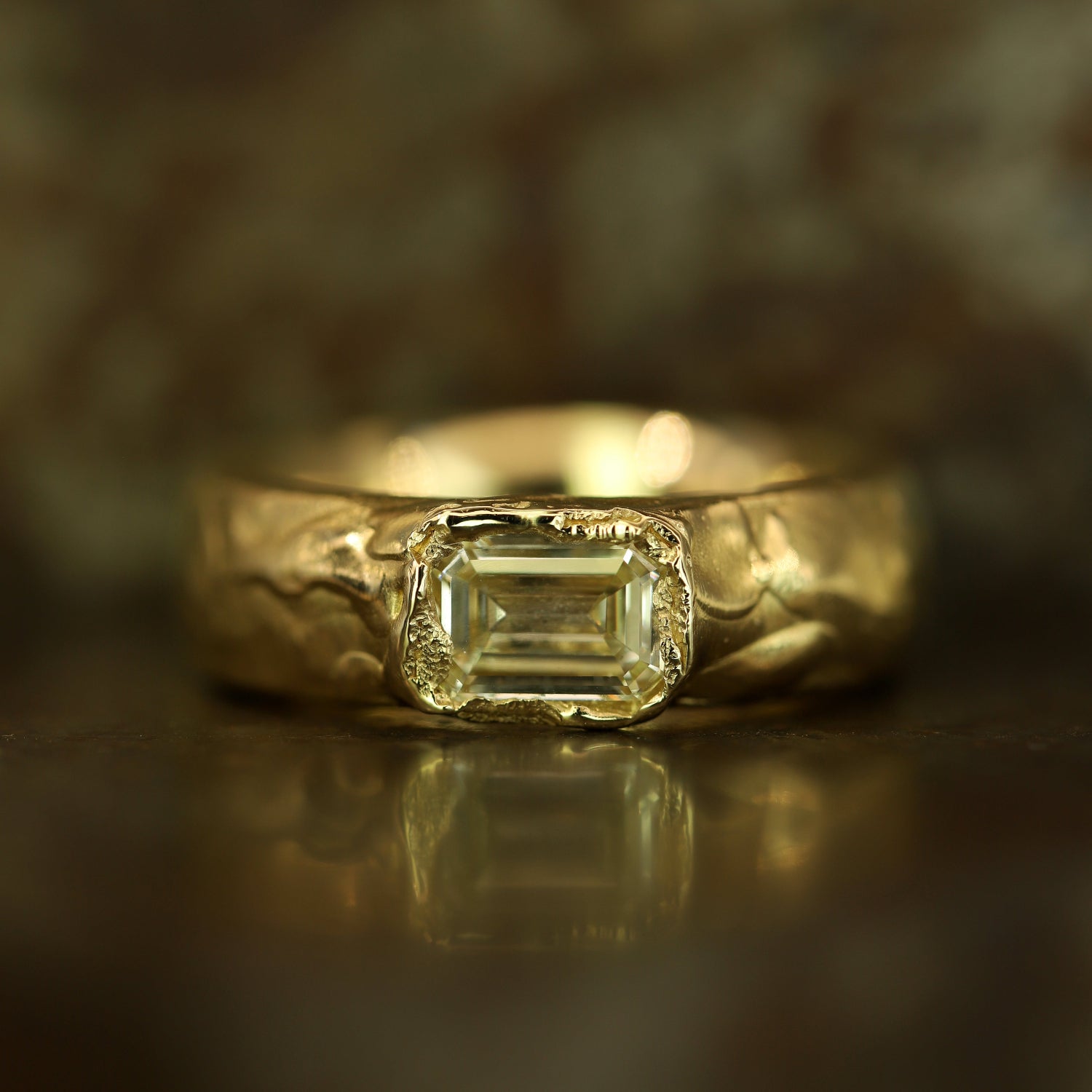 A gold ring featuring a rectangular emerald-cut clear gemstone, set against a dark, textured background with a soft reflection below.