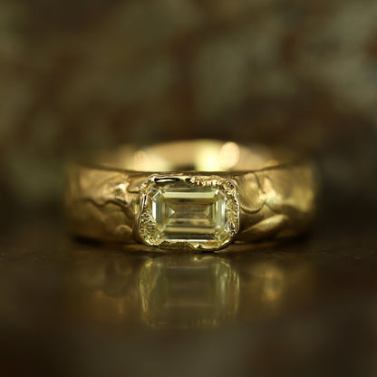 A gold ring featuring a rectangular emerald-cut clear gemstone, set against a dark, textured background with a soft reflection below.
