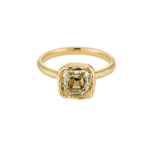 A gold ring featuring a square-cut, emerald-shaped clear gemstone in a textured bezel setting, shown on a plain white background.