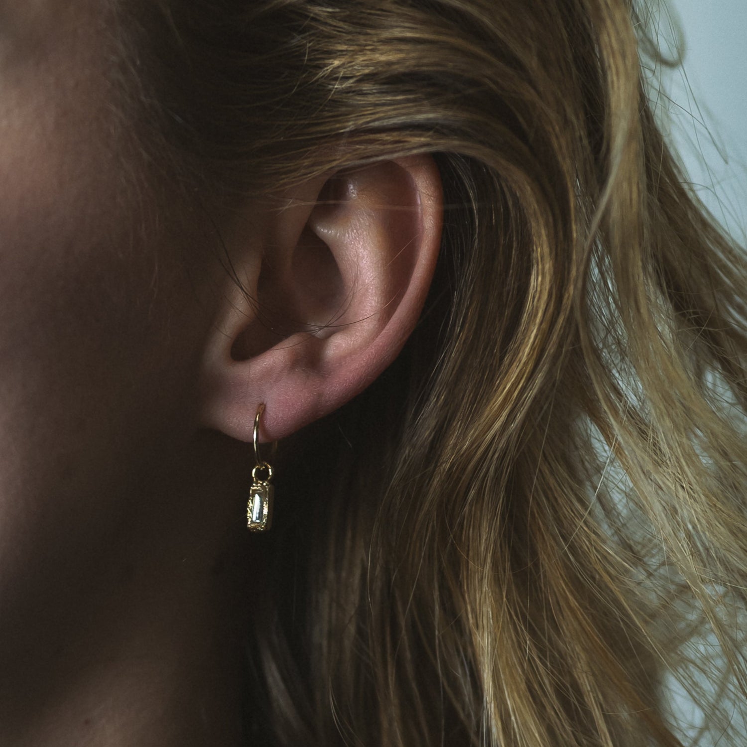 Ellis Mhairi Cameron - Close-up of an ear wearing a gold earring with a small charm, hair partially covering the face.