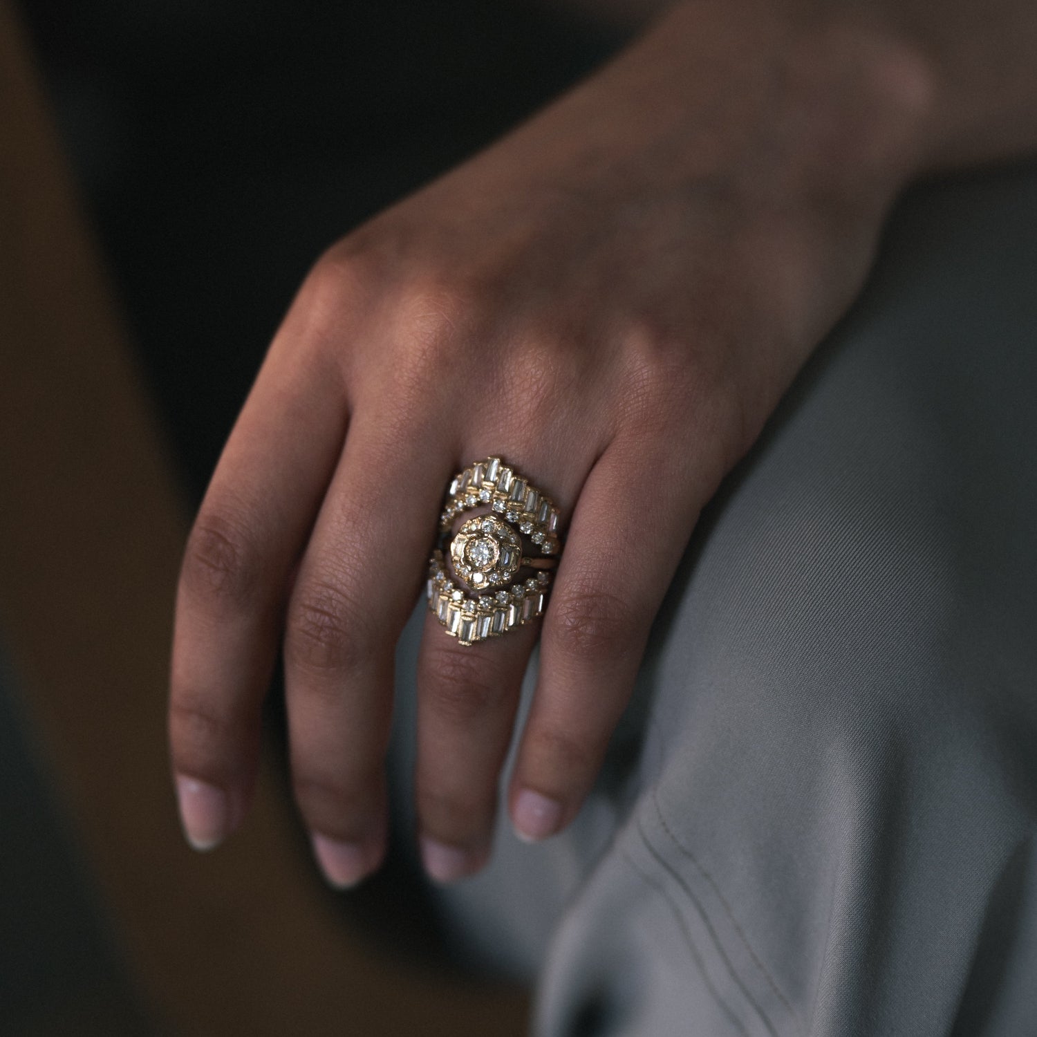 A close-up of a hand wearing a large, ornate gold ring with multiple clear gemstones, resting against light gray fabric.