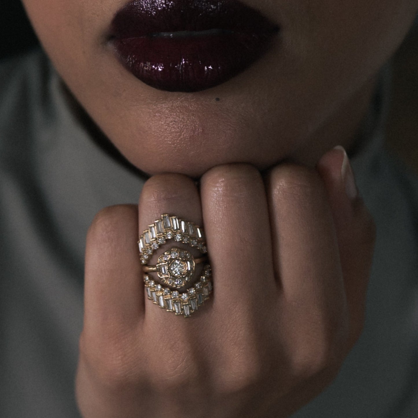 A close-up of a person's hand wearing a large, ornate ring with a central clear gemstone surrounded by multiple rows of small clear stones and gold-toned metal, set against a dark gray top and dark background.