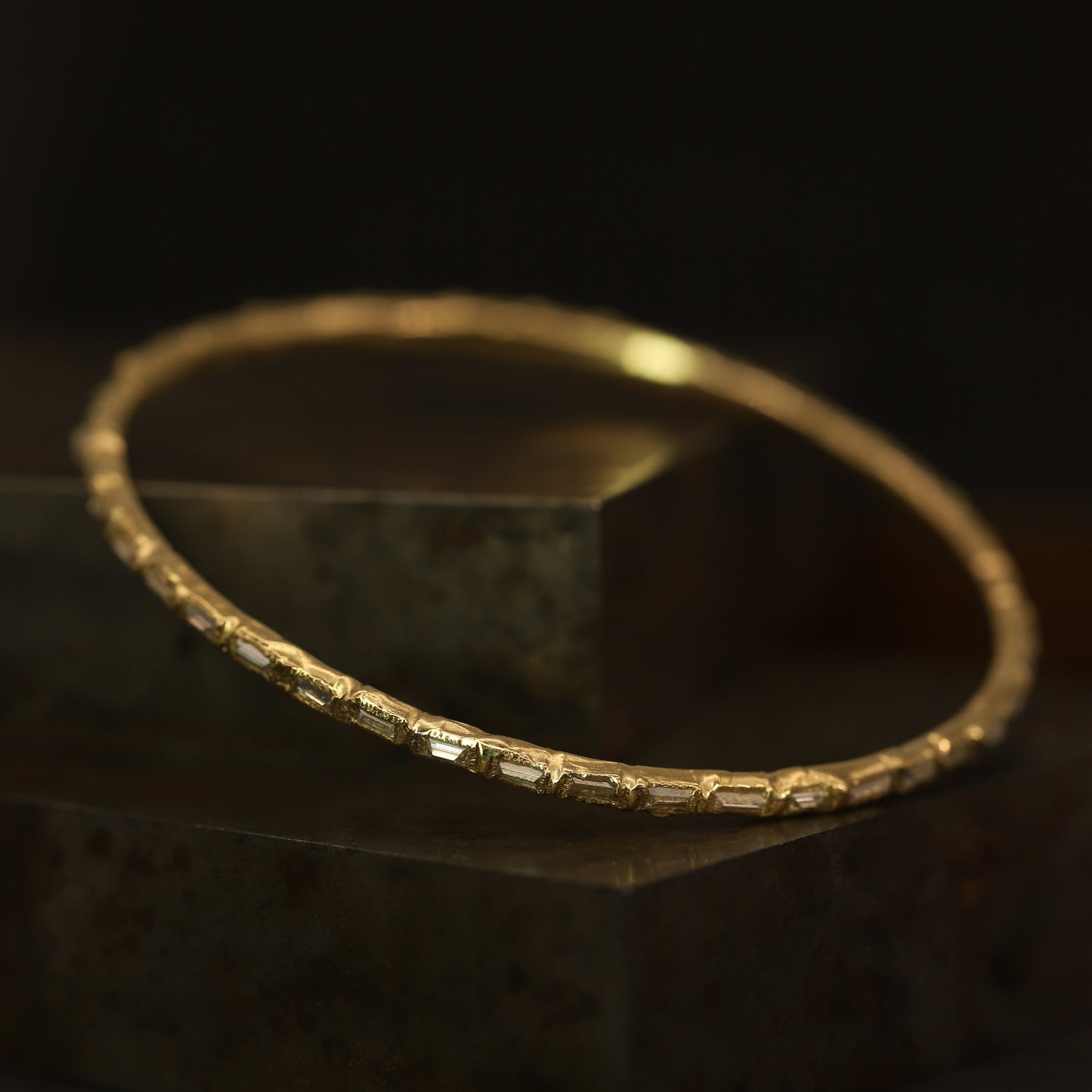 A gold bangle bracelet with baguette-cut clear stones, resting on a dark textured block against a black background.