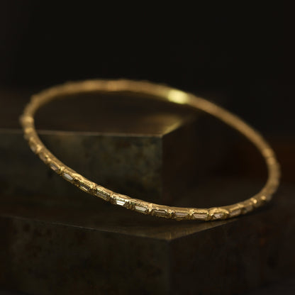 A gold bangle bracelet with baguette-cut clear stones, resting on a dark textured block against a black background.