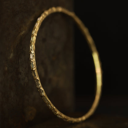 A textured gold bangle bracelet with a hammered, rustic finish, resting against a dark, rustic wooden background.