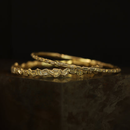 Two gold bangles with a textured, geometric segmented pattern, resting on a dark reflective surface against a black background.