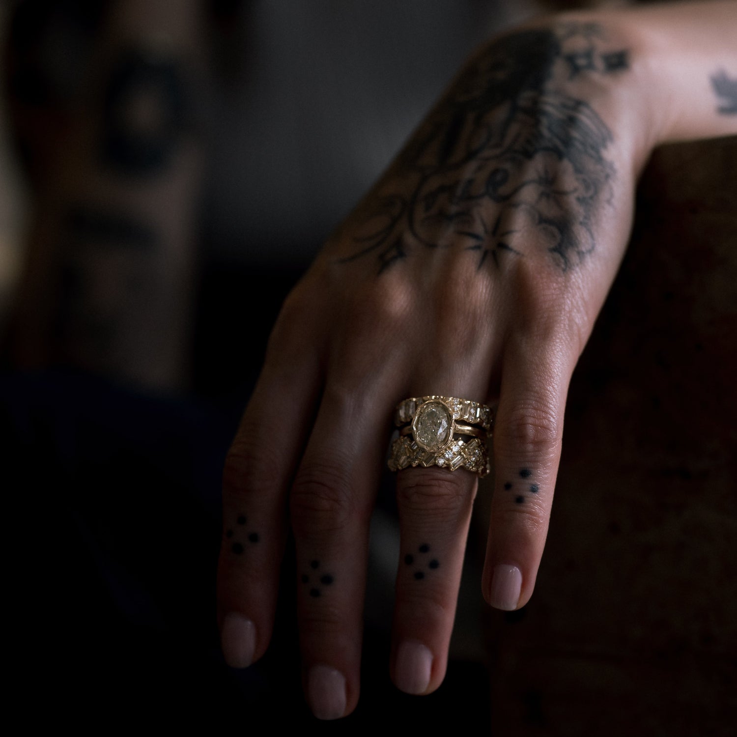 Ellis Mhairi Cameron - Hand with tattoos wearing a gold ring against a dark background