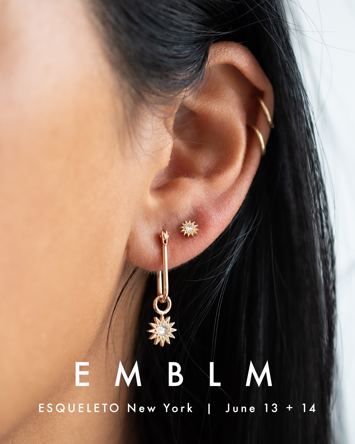 Close-up of an ear wearing gold earrings with 'EMBLM' branding.