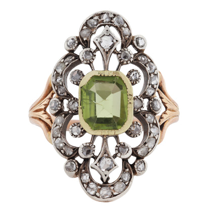 An ornate antique ring featuring a central green peridot gemstone surrounded by a silver filigree setting with small diamond stones, and gold side panels on a white background.