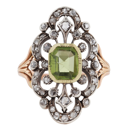 An ornate antique ring featuring a central green peridot gemstone surrounded by a silver filigree setting with small diamond stones, and gold side panels on a white background.