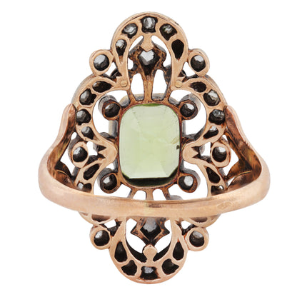 A rose gold filigree ring featuring a central green peridot gemstone and small dark gemstone accents, shown on a plain white background.