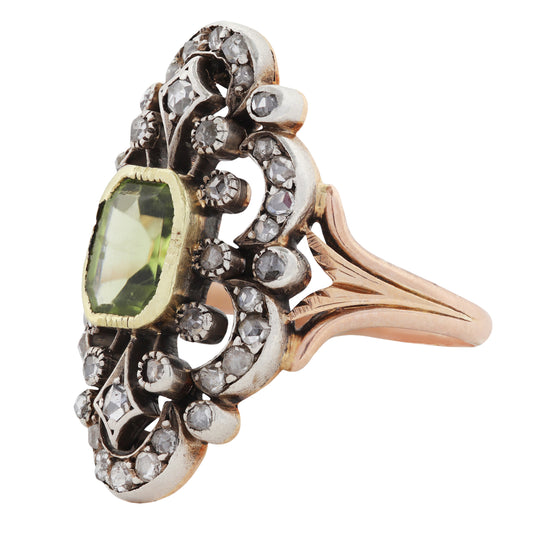 An ornate antique ring featuring a central green peridot gemstone surrounded by a silver filigree setting with small diamond stones, and gold side panels on a white background.
