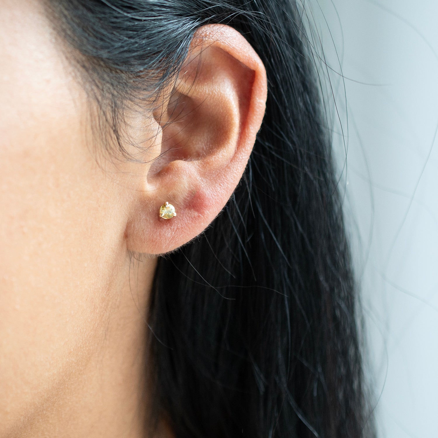 A close-up of a person's ear wearing a small gold stud earring, with dark hair and a light gray background.