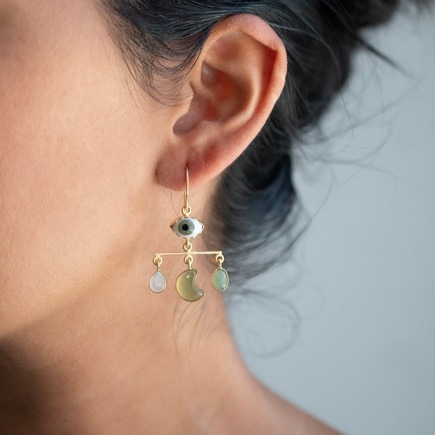 A close-up of a person's ear wearing a delicate gold dangle earring with an evil eye charm and three dangling charms, including a crescent moon and oval stones, against a soft gray background.