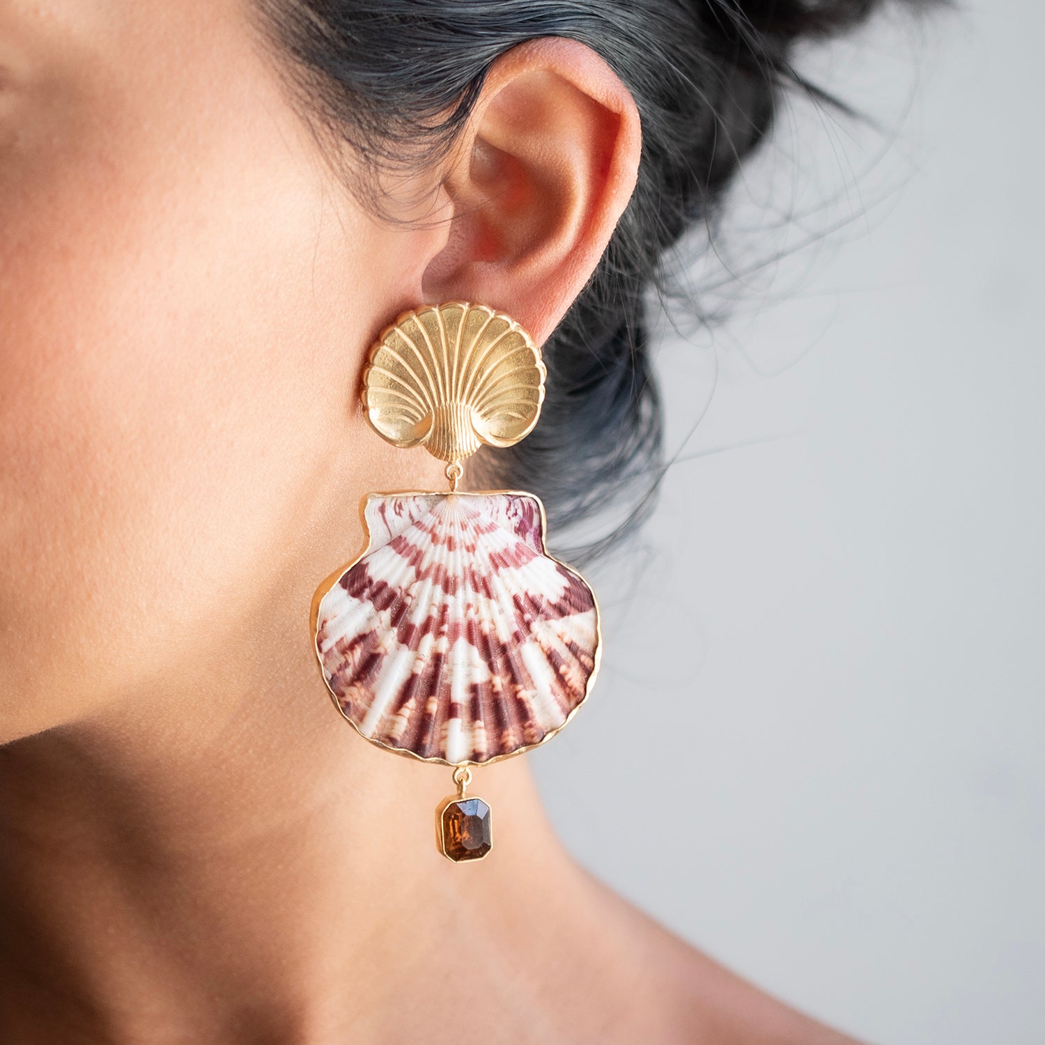A close-up of a person's ear wearing a large shell-shaped drop earring with a gold scallop stud, a pink and white striped scallop shell, and a small dangling amber gemstone, against a light gray background.