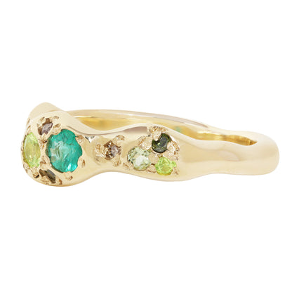 A gold ring with a wavy, organic shape, featuring a large round green emerald and smaller green and brown stones, set against a plain white background.