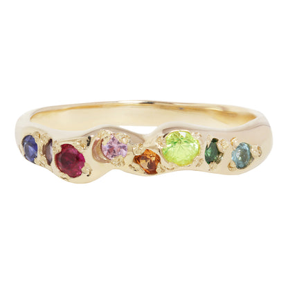 A gold band ring featuring a wavy channel setting with multiple round-cut gemstones in various colors including blue, purple, red, orange, green, and light blue, set against a plain white background.