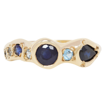 A gold ring featuring a wavy channel setting with a large oval dark blue sapphire in the center, flanked by smaller round blue and light blue round-cut gemstones, set against a plain white background.