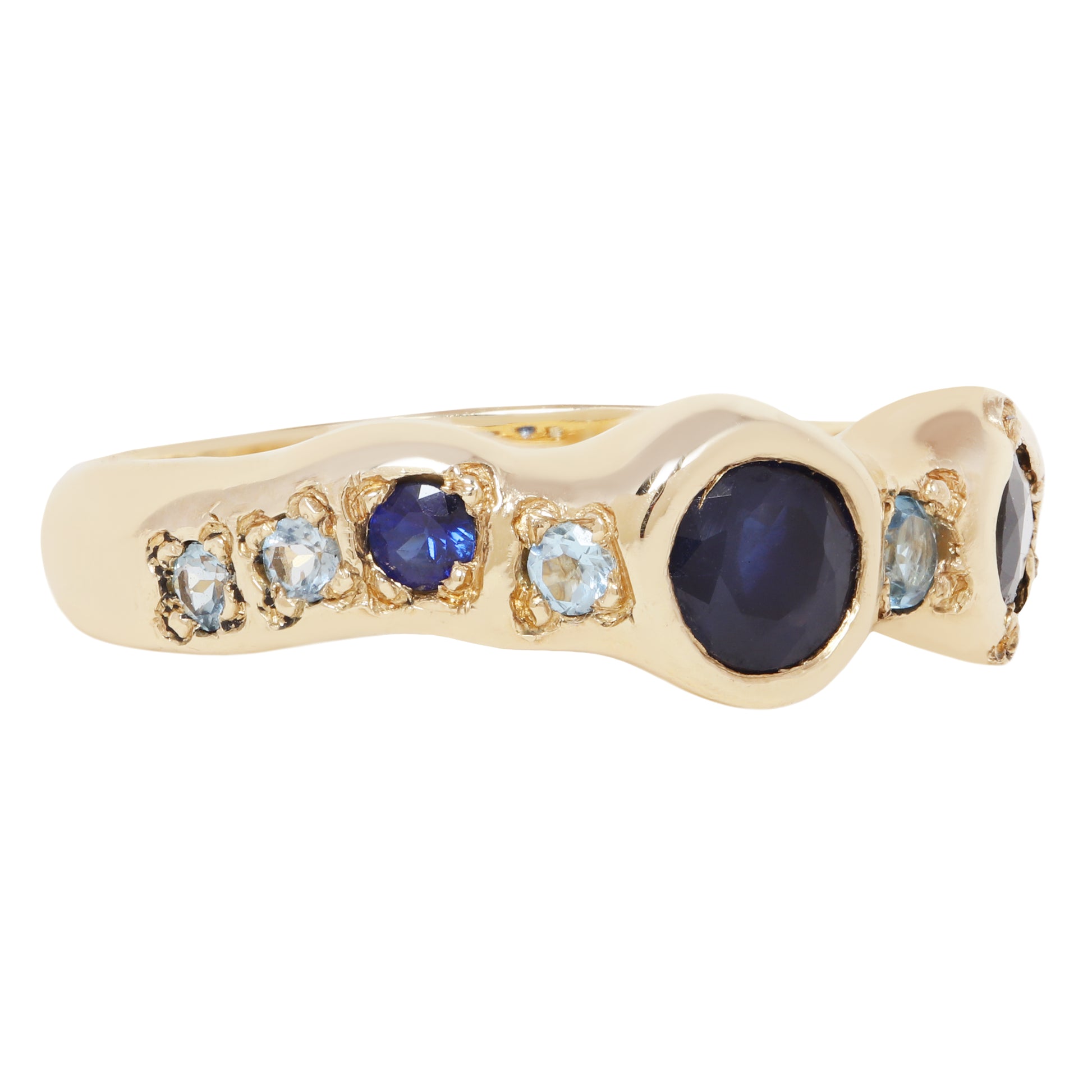A gold ring featuring a wavy, organic-shaped setting with a large oval dark blue cabochon center stone, flanked by smaller round blue and light blue gemstones in gold bezels, shown on a plain white background.