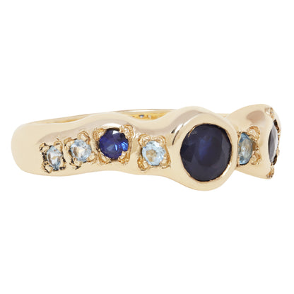 A gold ring featuring a wavy, organic-shaped setting with a large oval dark blue cabochon center stone, flanked by smaller round blue and light blue gemstones in gold bezels, shown on a plain white background.