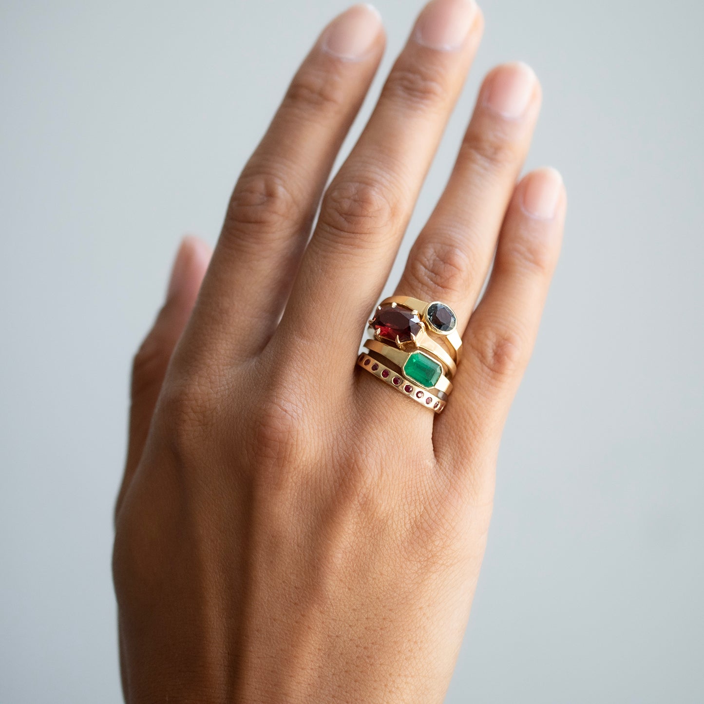 A gold ring with multiple colorful gemstones, including green, red, and black, worn on a person's hand against a light gray background.