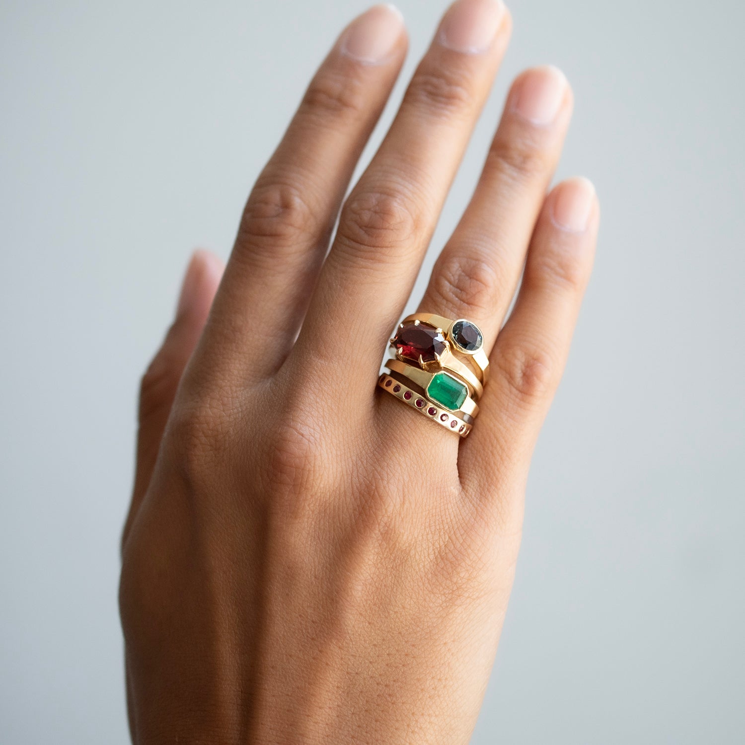 A gold ring with multiple colorful gemstones, including green, red, and black, worn on a person's hand against a light gray background.