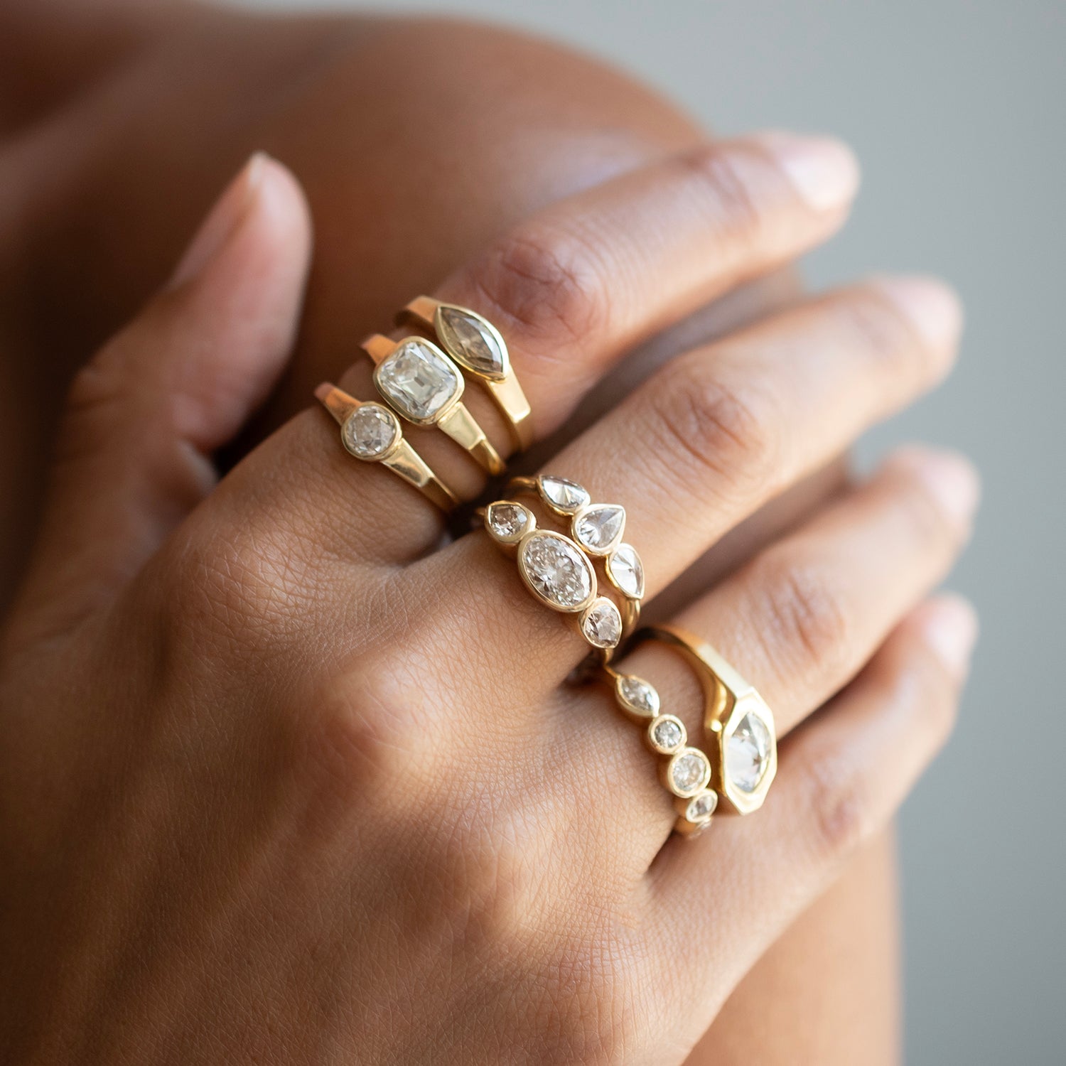 A close-up of a hand wearing multiple gold rings with clear gemstones in various shapes, set against a soft gray background.