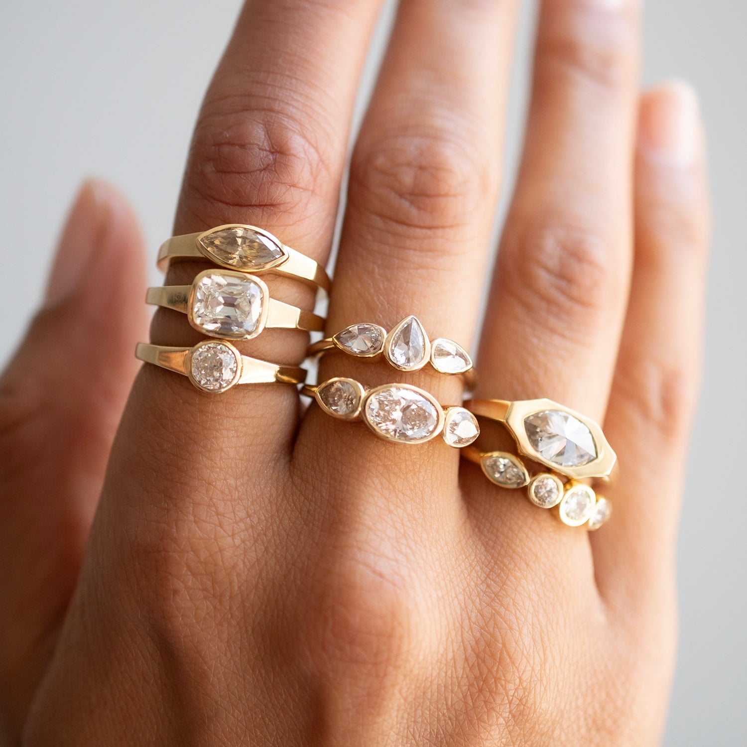 A close-up of a hand wearing multiple gold rings with clear gemstones in marquise, round, and oval settings against a soft light gray background.