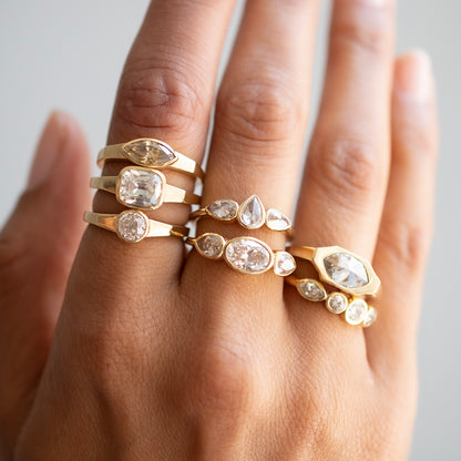 A close-up of a hand wearing multiple gold rings with clear gemstones in marquise, round, and oval settings against a soft light gray background.