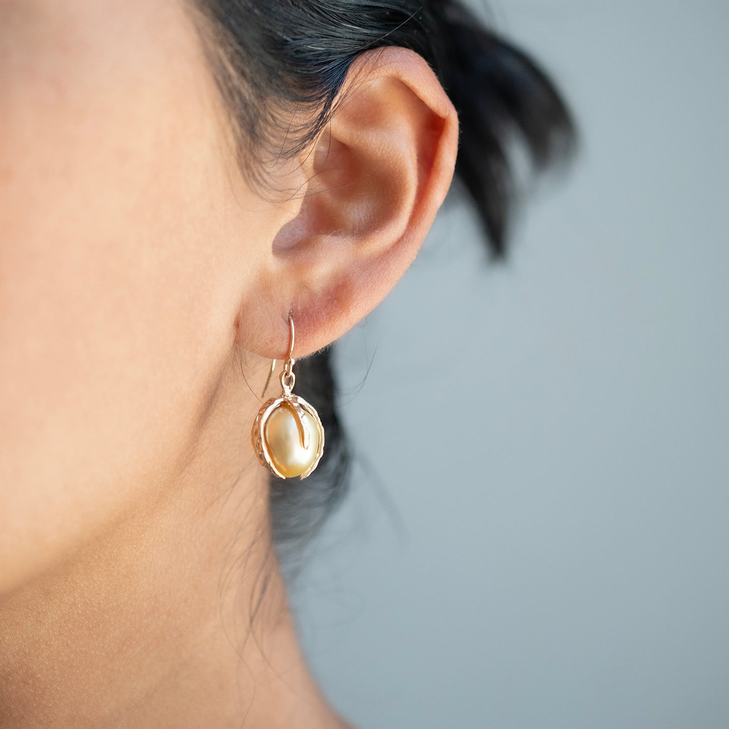 A close-up of a person's ear wearing a delicate gold hoop earring with a large, irregular gold baroque pearl dangle.