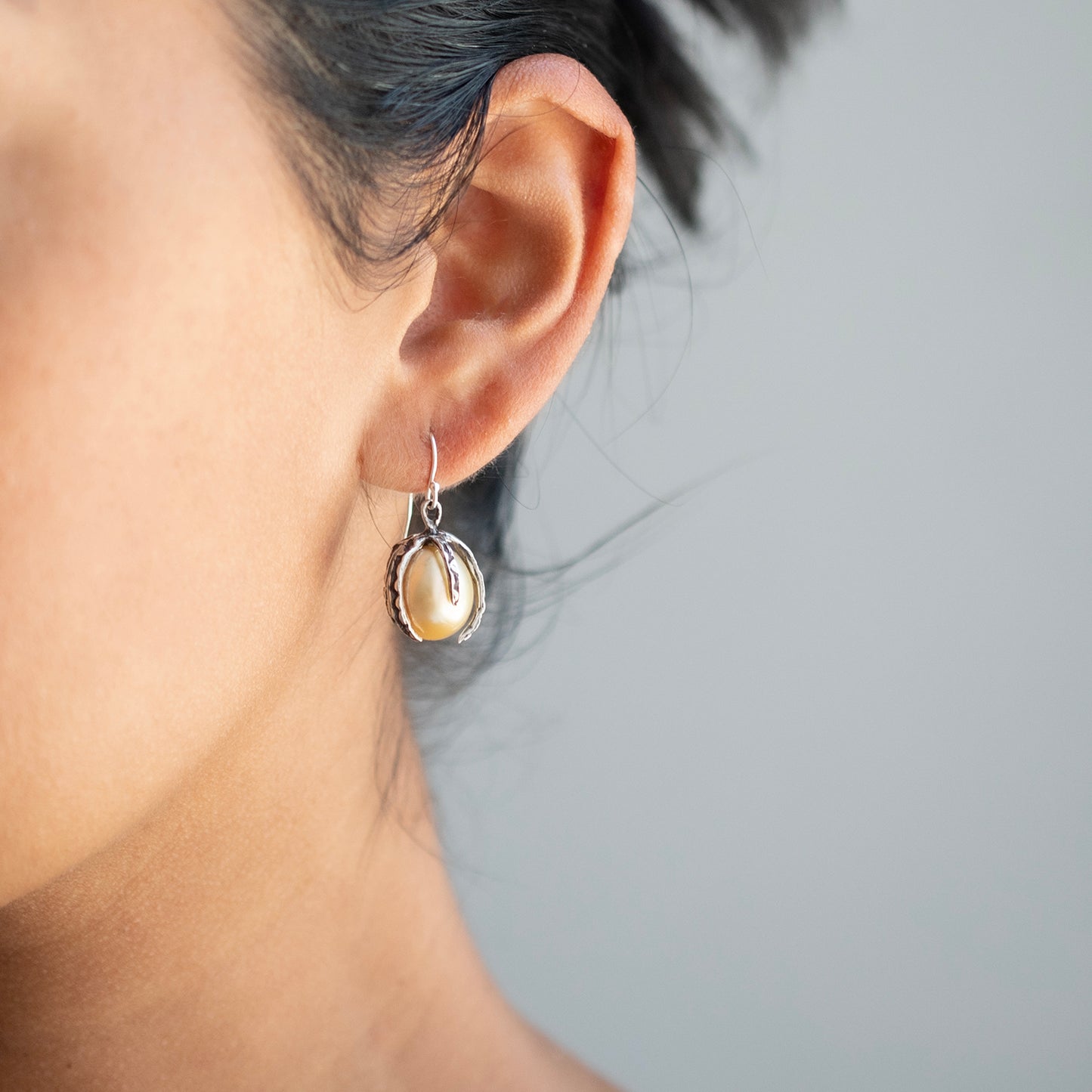 A close-up of a person's ear wearing a small silver hoop earring with a dangling irregular golden pearl.