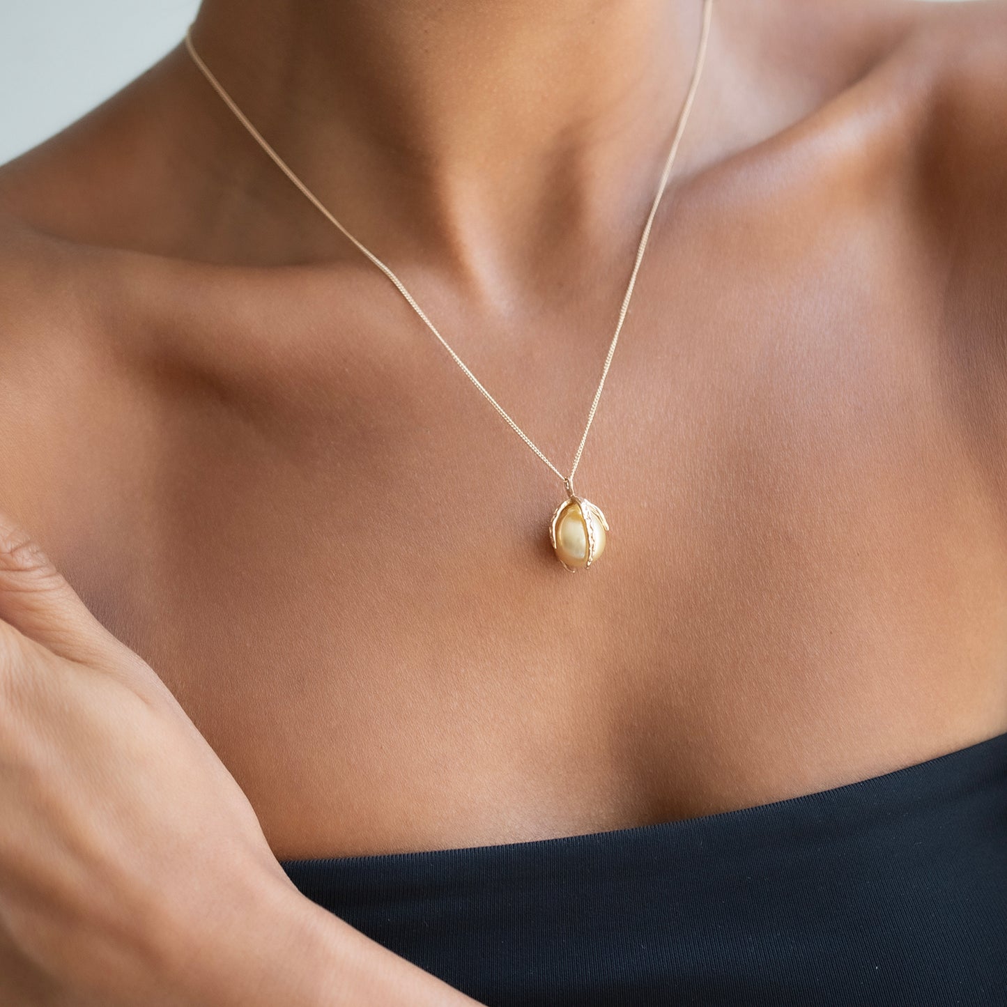 A close-up of a model's chest wearing a delicate gold chain necklace with a pearl, textured gold pendant.