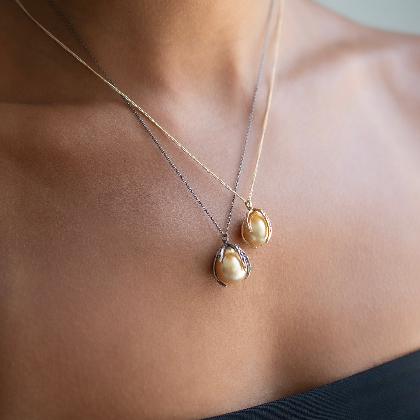 Two delicate necklaces worn on a model's neck, featuring a larger gold pendant with a baroque pearl and a smaller silver pendant with a cream pearl.