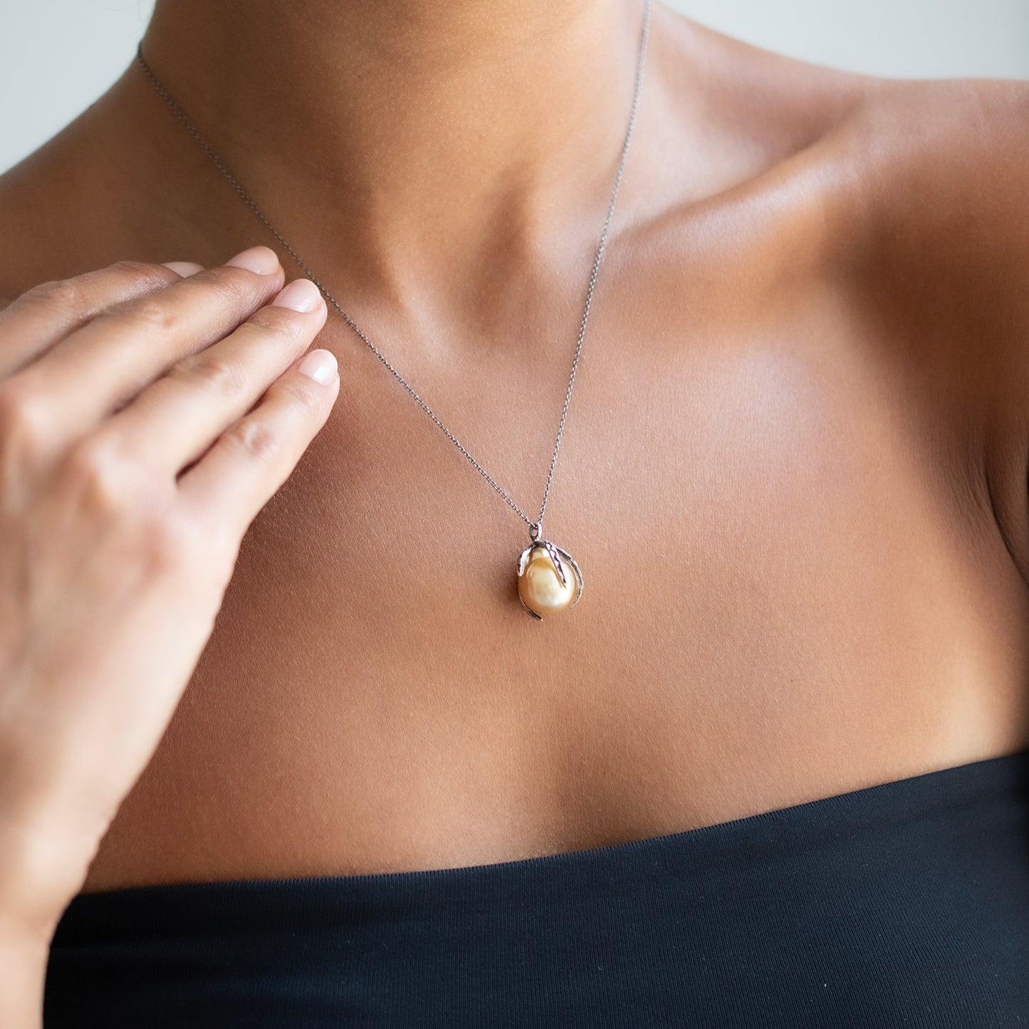 A close-up of a person wearing a delicate silver chain necklace with a single round, cream-colored pearl pendant, styled with a black strapless top.
