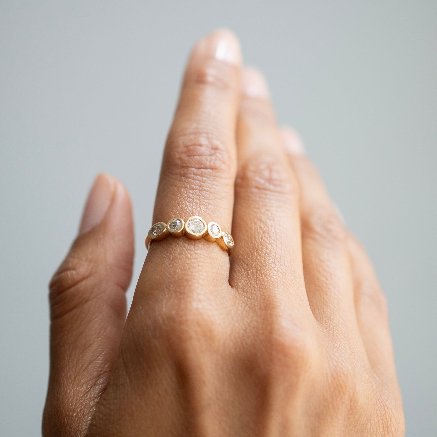 A close-up of a hand wearing a delicate gold band ring adorned with small round clear stones, set against a soft gray background.